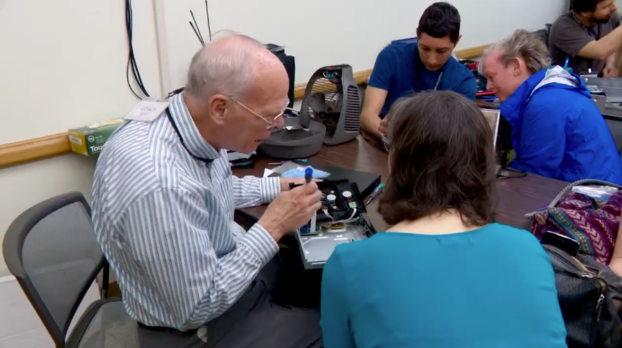 Man fixing an item sitting in a room with other people. 