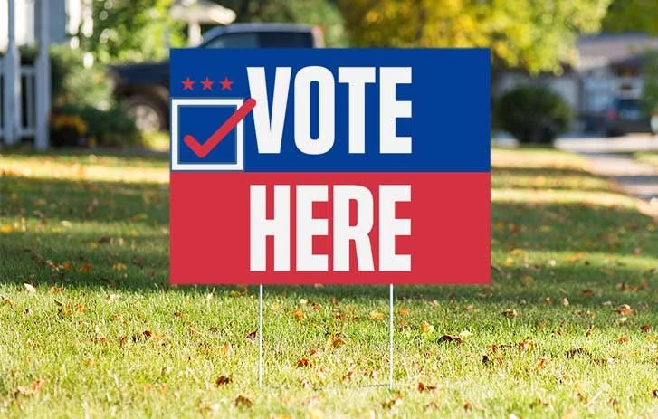 And election sign that says vote here stands on a lawn.
