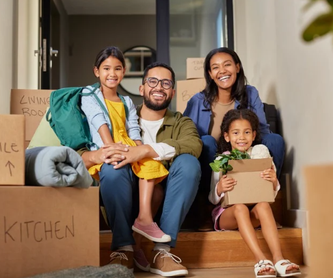 A family sits on steps of their home, smiling, with moving boxes around them.
