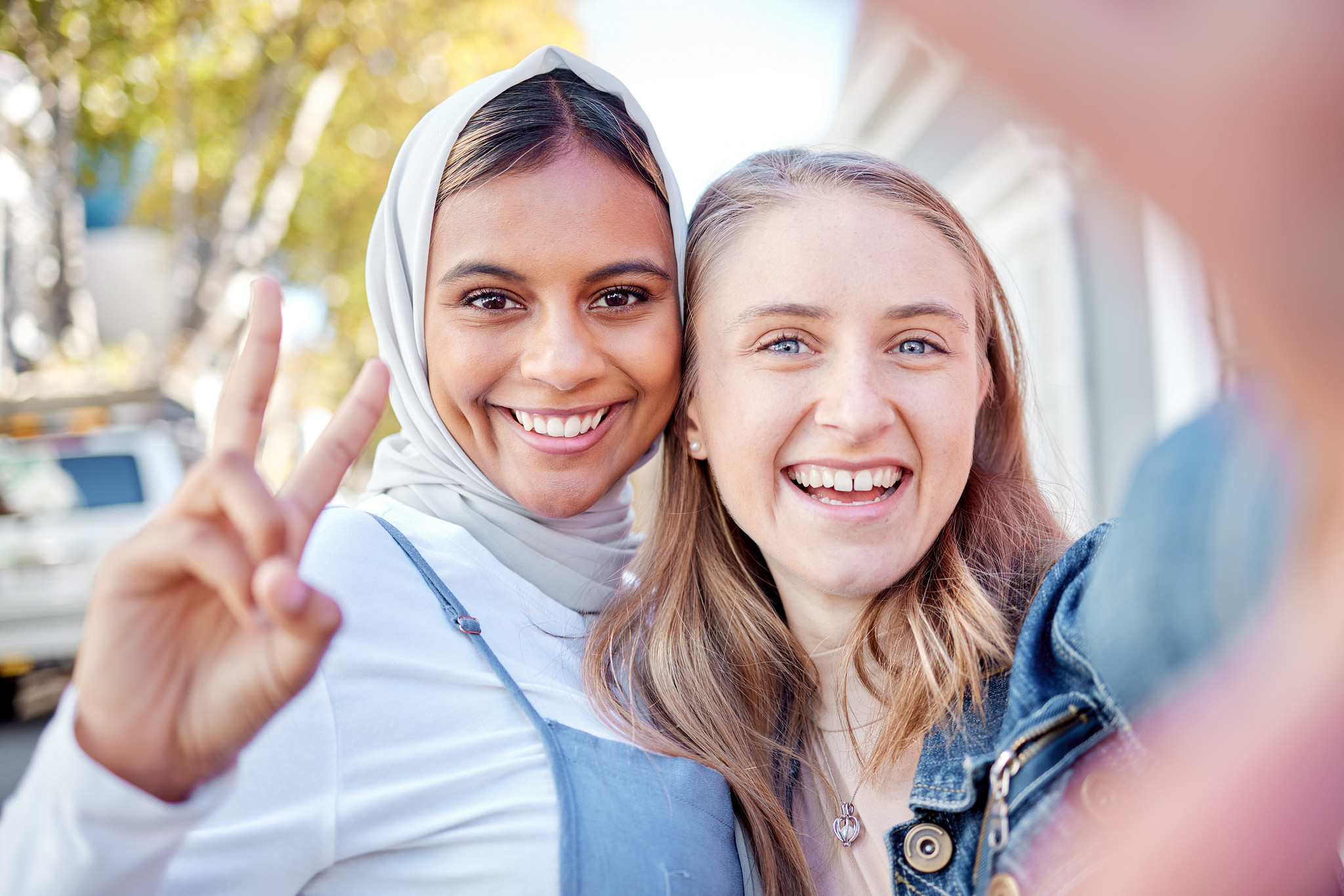 Blonde white woman and a woman wearing a hijab flash a peace sign at camera posing for photo. 