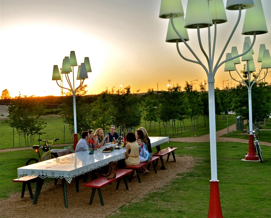 Six people gathered around a long white outdoor table with red benches in a park at sunset, enjoying food and drinks beneath whimsical tree-like lamp posts, with bicycles nearby and a serene landscape of grass, paths, and young trees in the background.