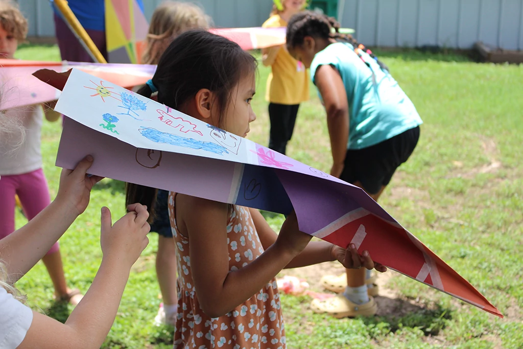 A group of youth students outside on the grass in the sunshine ready themselves to test race their giant paper airplanes made of many different colored sheets of construction paper taped together and covered in drawings