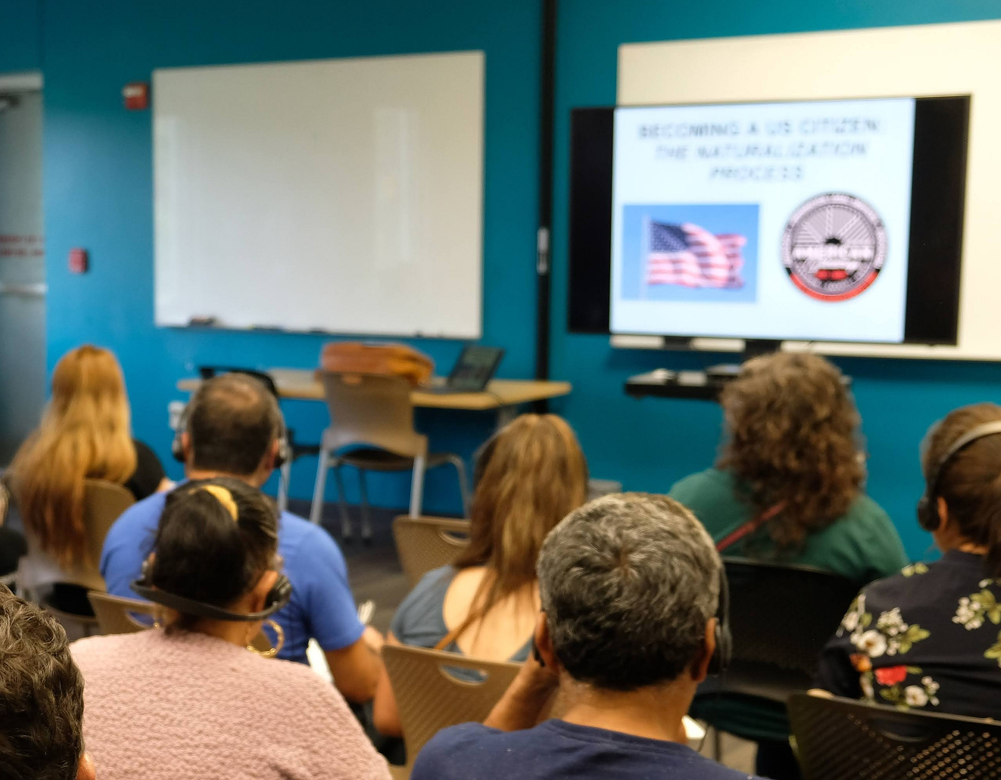 Back angle of a group of people sitting and facing a presentation. Some people are wearing interpretation equipment.
