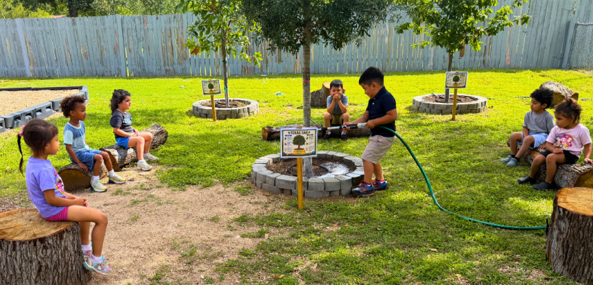 A student waters a tree with a hose while other students sit on logs looking on.