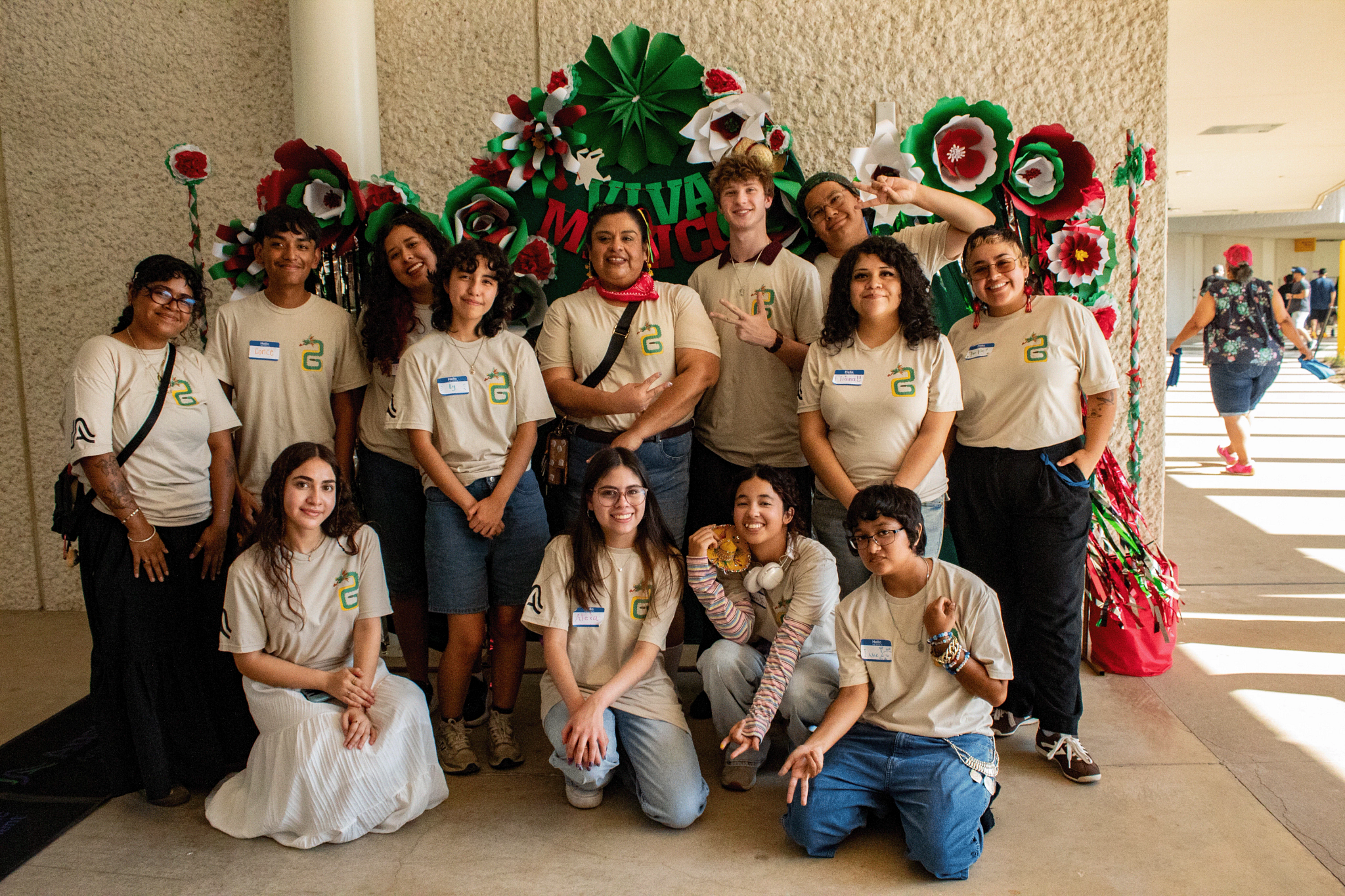 A group of teenagers and one adult smile and stand before a green backdrop with red white and green flowers