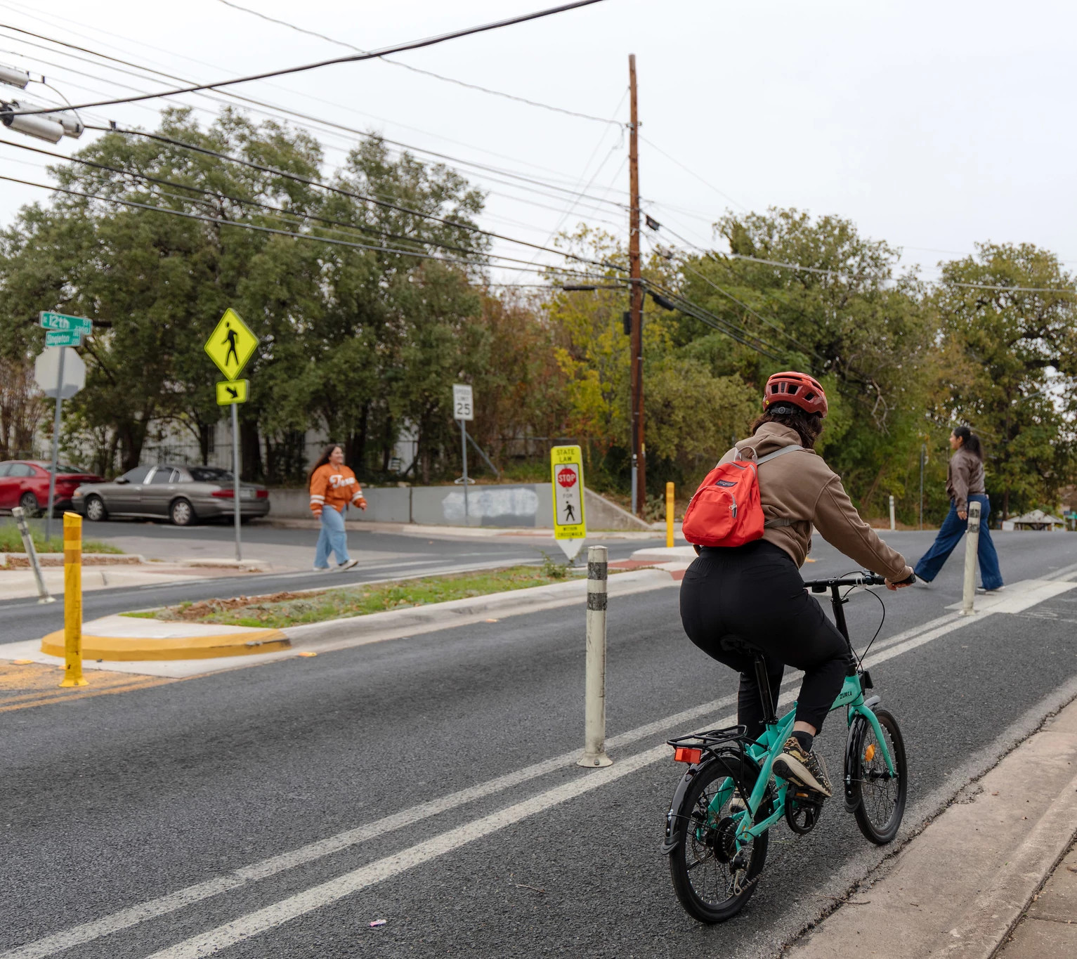 A person rides a bicycle in a protected bike lane while people cross at a crosswalk with a pedestrian crossing island on East 12th Street.