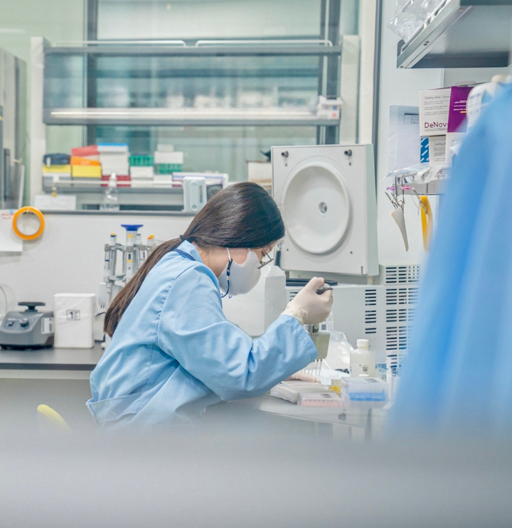 Woman wearing a lab coat, glasses, gloves, and a mask leans over her work in a genomics lab