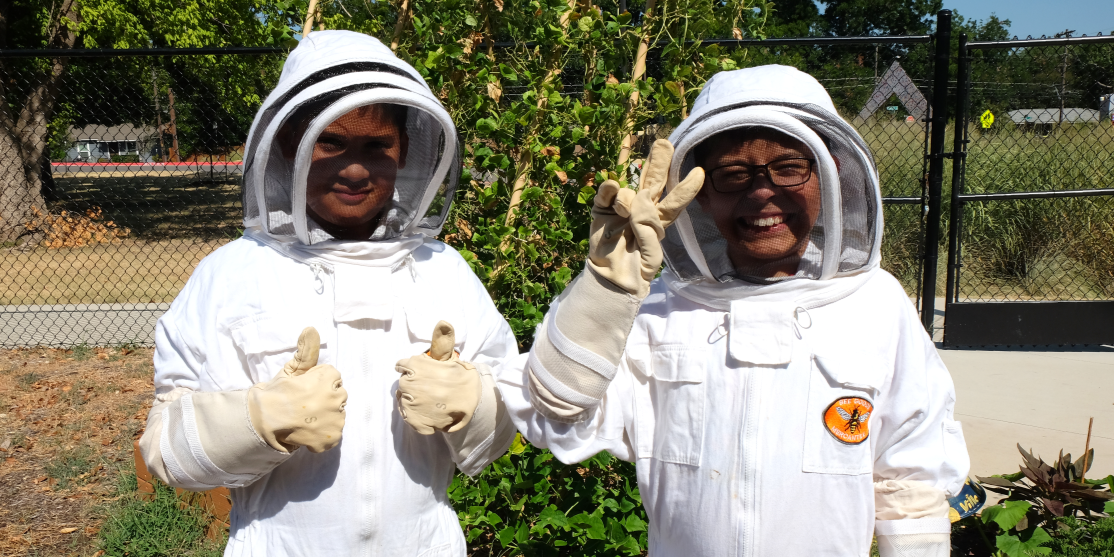 Two students in beekeeper suits. One is giving a thumbs up. The other is giving a peace sign.