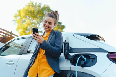 A woman reads from her phone while leaning against her electric vehicle which is plugged in and charging