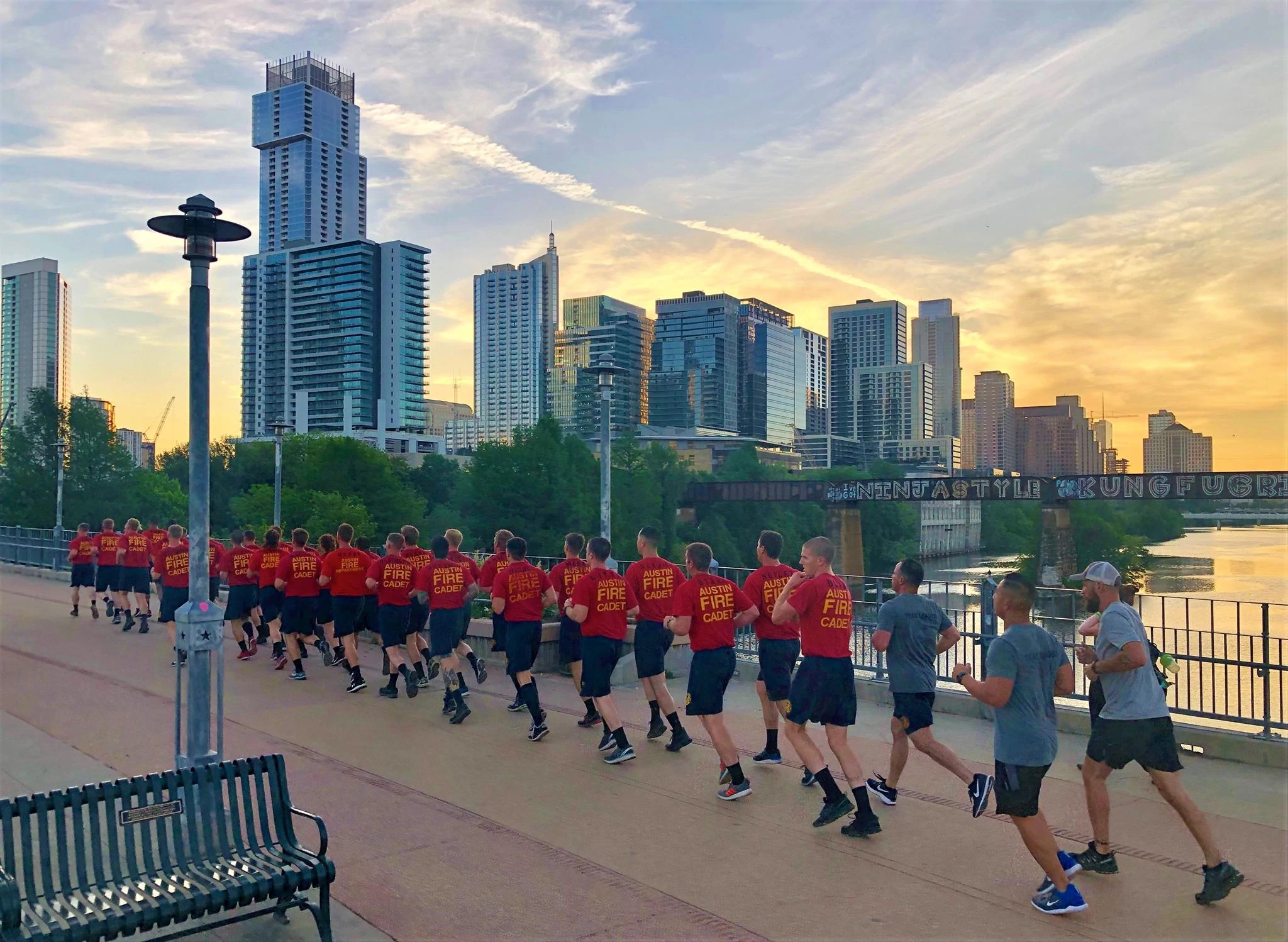 Group of Austin Fire cadets running on a bridge across Ladybird Lake with the sun rising behind the Austin skyline in the background