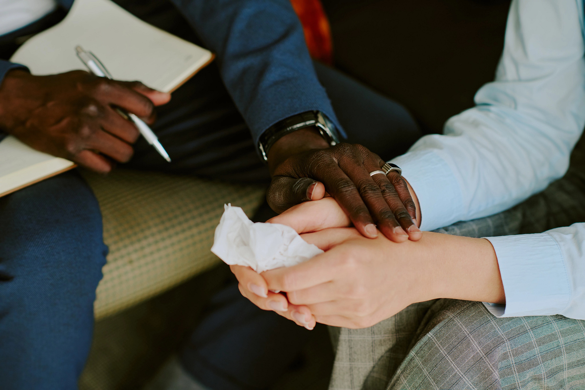 tight photo of a black man's and white woman's hands. The man holds a notepad and pen in one hand and holds the woman's hands with the other. She holds a facial tissue.