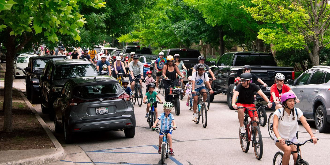 A group of people of all ages ride their bikes down a tree-lined street.