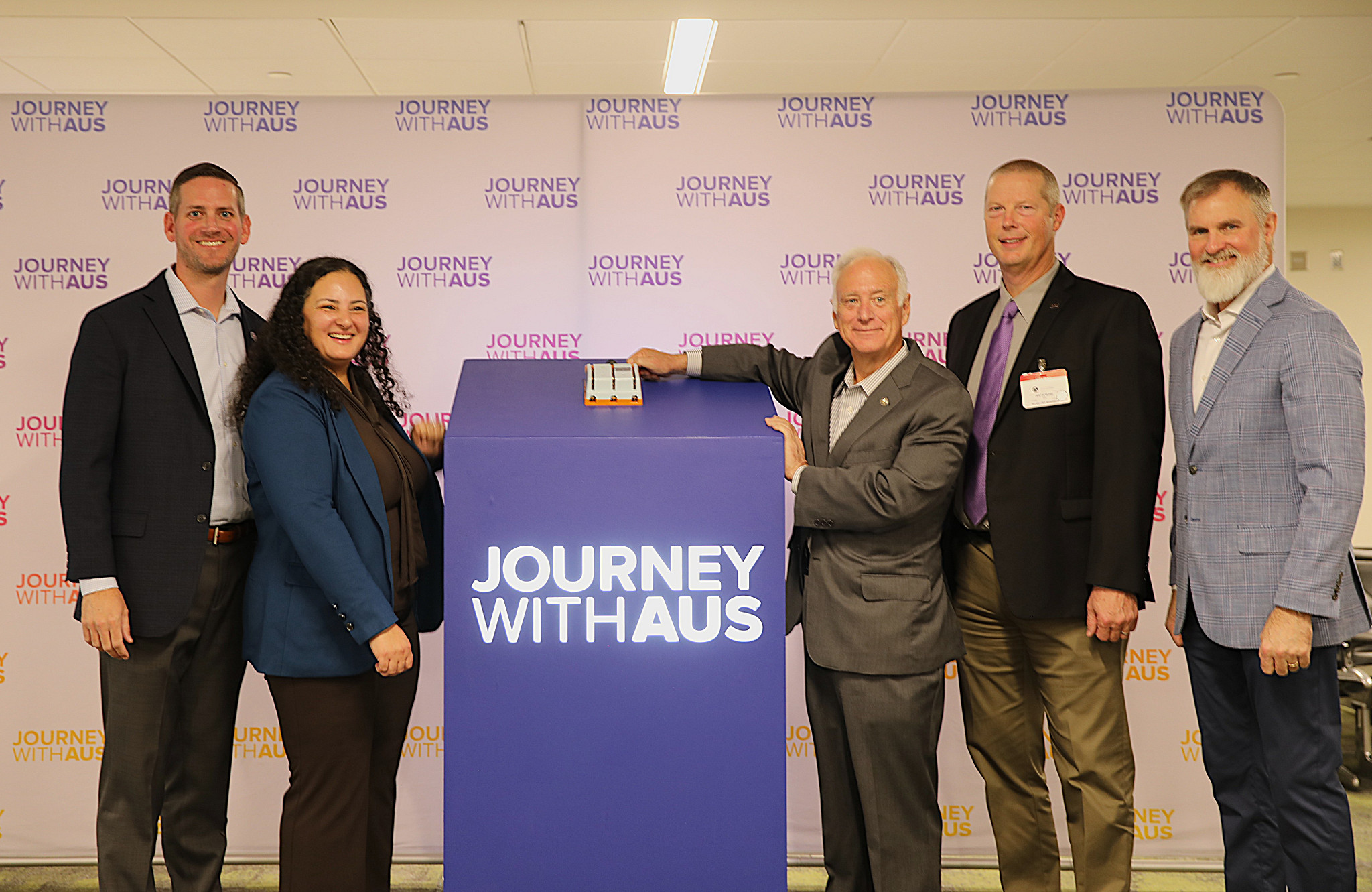 A group of five adults stands indoors in front of a backdrop that displays the repeated words JOURNEY WITH AUS. A large rectangular podium with the same words is positioned in the center. One adult is touching a small rectangular object placed on top of t