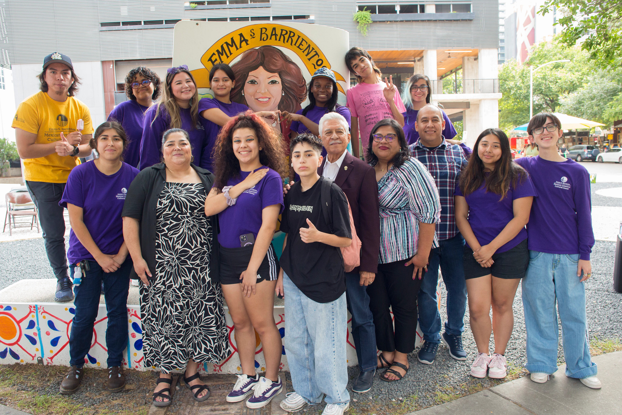Group of teens and adults smile for a picture in front of a mural of a woman that says Emma S. Barrientos