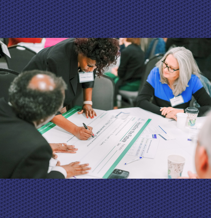A Black man, Black woman, and white woman together at a table for a workshop. The Black woman is standing and writing on chart paper. 