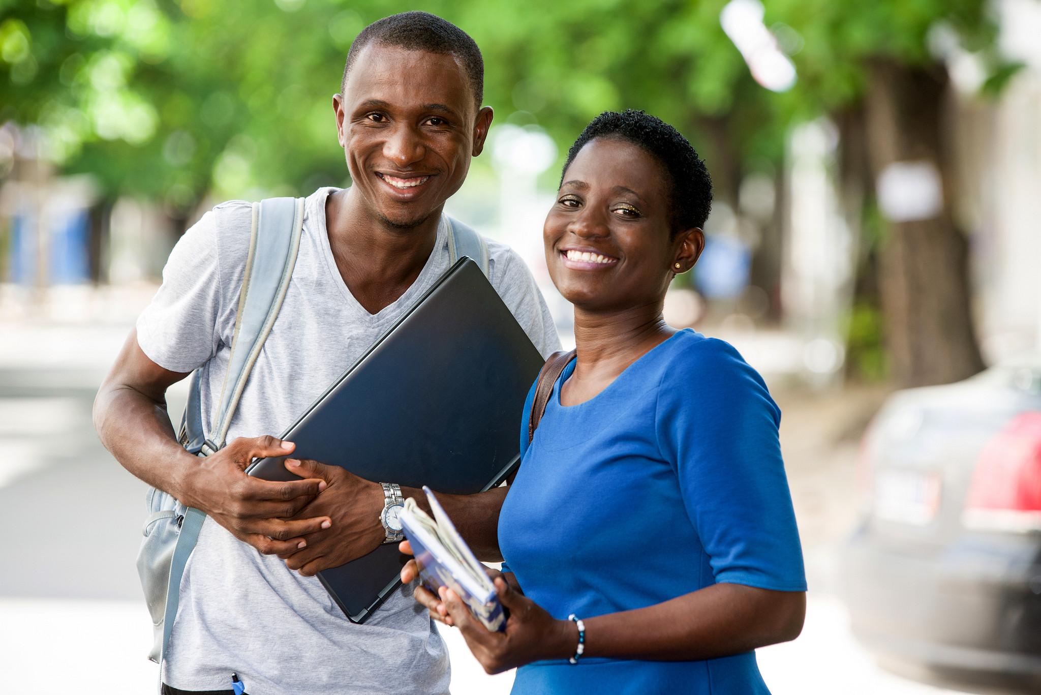 A black man and woman smile at the camera while holding a laptop, books, and wearing a backpack.