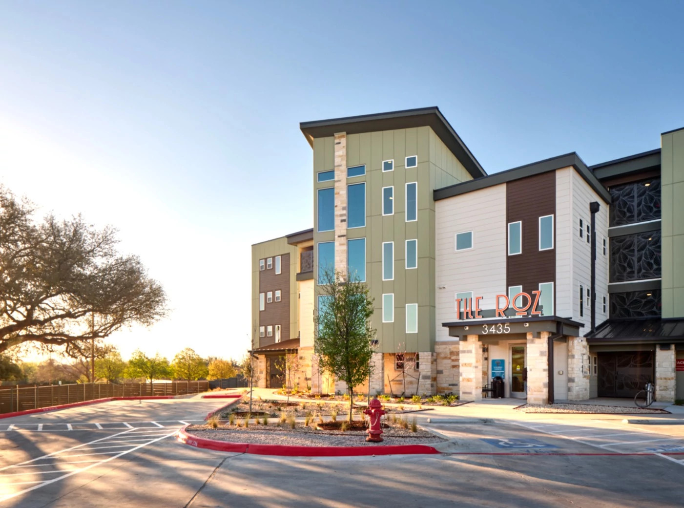 Image of the newly constructed permanent supportive housing community called The Roz. The image shows multiple multi-story buildings of green, beige and brown. The first floor is faced in white stone.