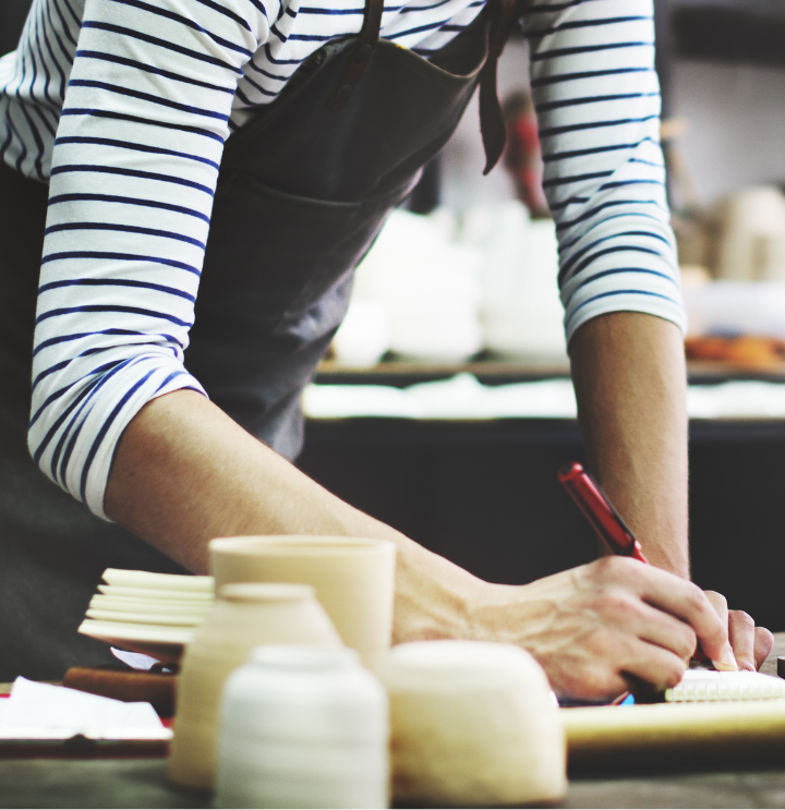 individual in a work apron writing in a notepad on their desk which is covered in pottery