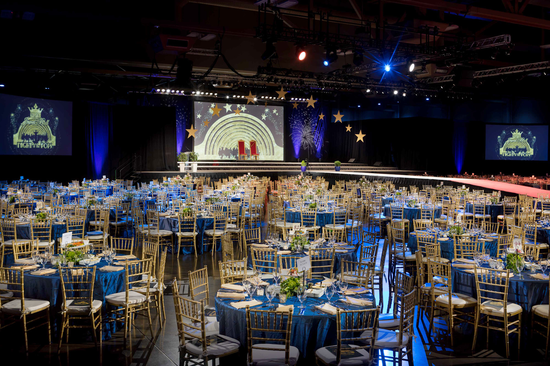 Image of a Palmer Events Center exhibit hall during the Jewel Ball filled with dinner tables with blue table cloths, flowers, and table settings with a stage in the background