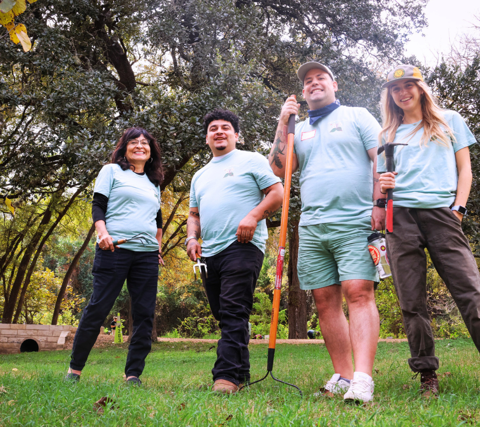 A diverse group of people stand in a greenspace in Tree Ambassador t-shirts. They smile and hold landscaping tools.