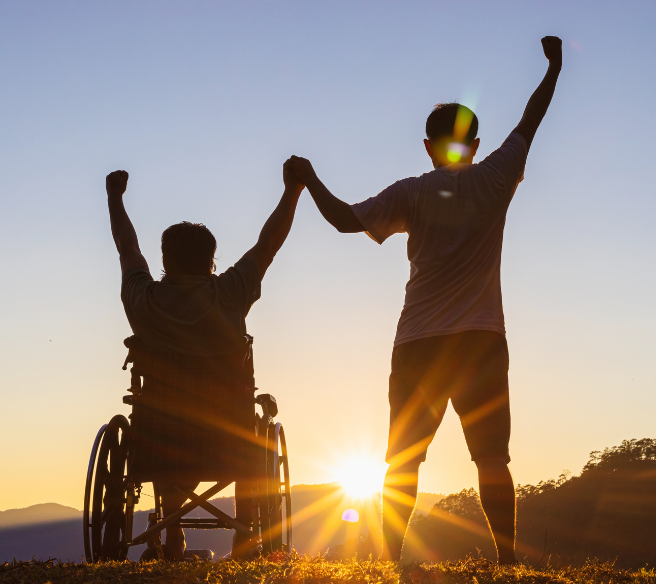 two men, one standing and one in a wheelchair, stand triumphantly on a hill in front of a sunset.