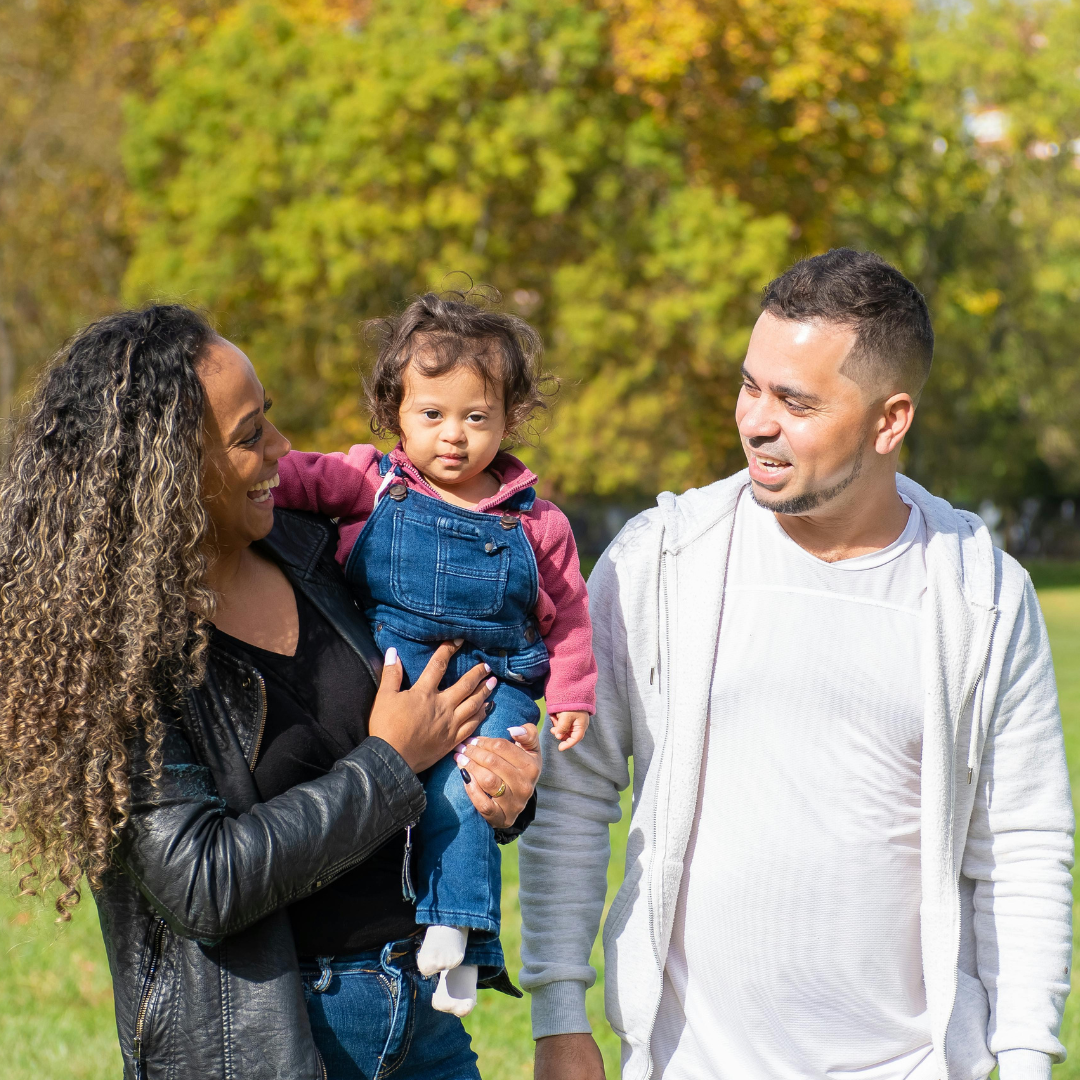 A multiracial family walks in a park with green field behind then. A woman of color with curly hair holds a toddler of color. A man of color smiles looking at the small girl.