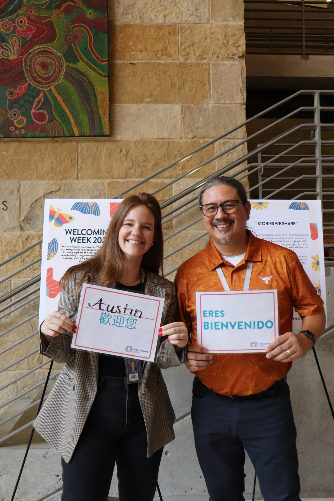 A white woman with straight brown hair holds a sign saying Austin Welcomes You in Chinese and is smiling. Next to her is a man of color smiling wearing glasses and a UT orange shirt holding a sign that says Austin Welcomes You in Spanish.