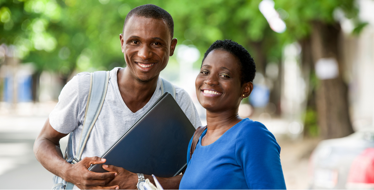 A Black man with a backpack on and holding a laptop and a Black woman holding a notebook smile wide at the camera. 