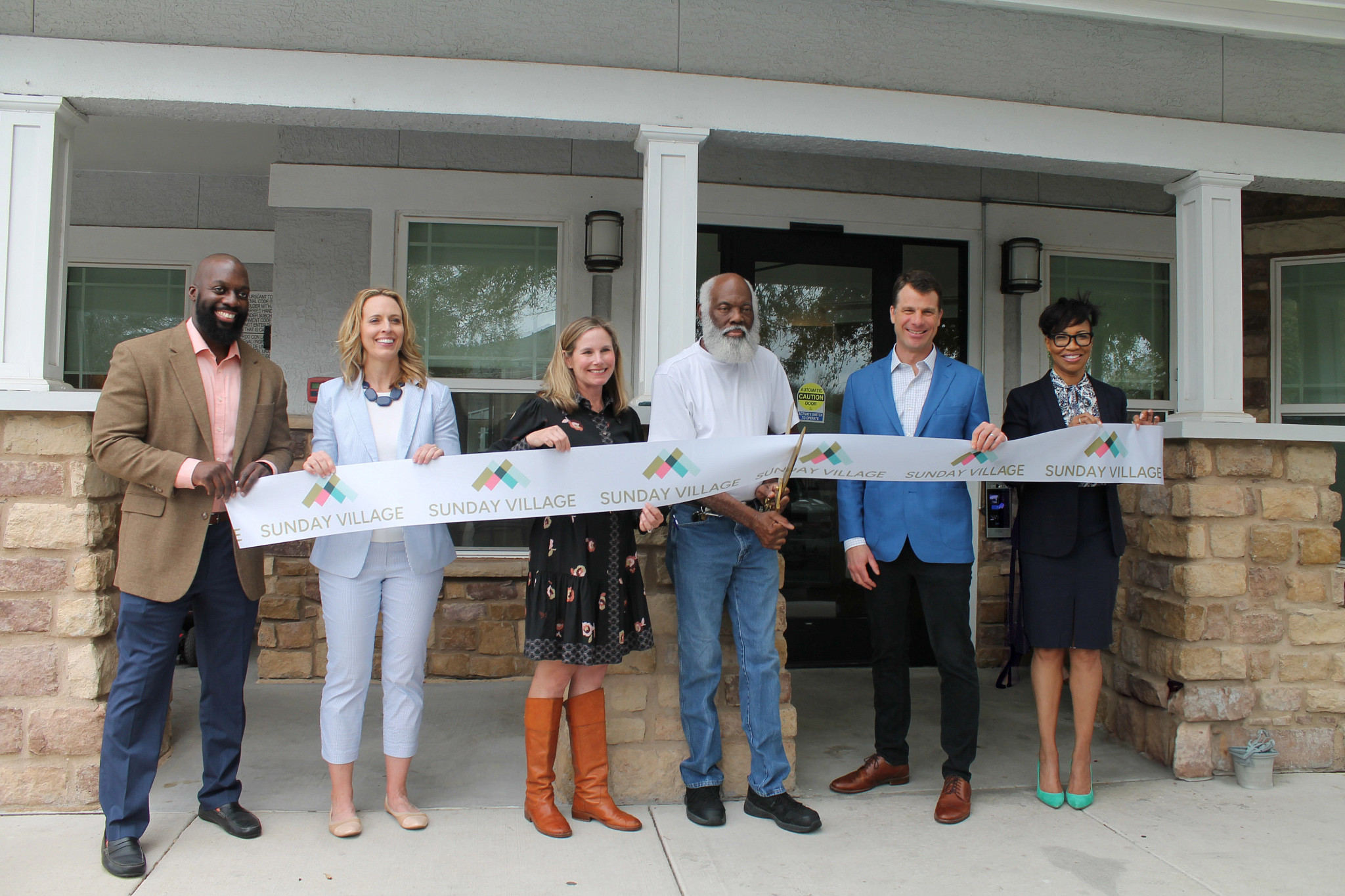 Image shows five individuals holding a long white ribbon with the repeating words Sunday Village. A man in the center holds large gold scissors and is cutting the ribbon.