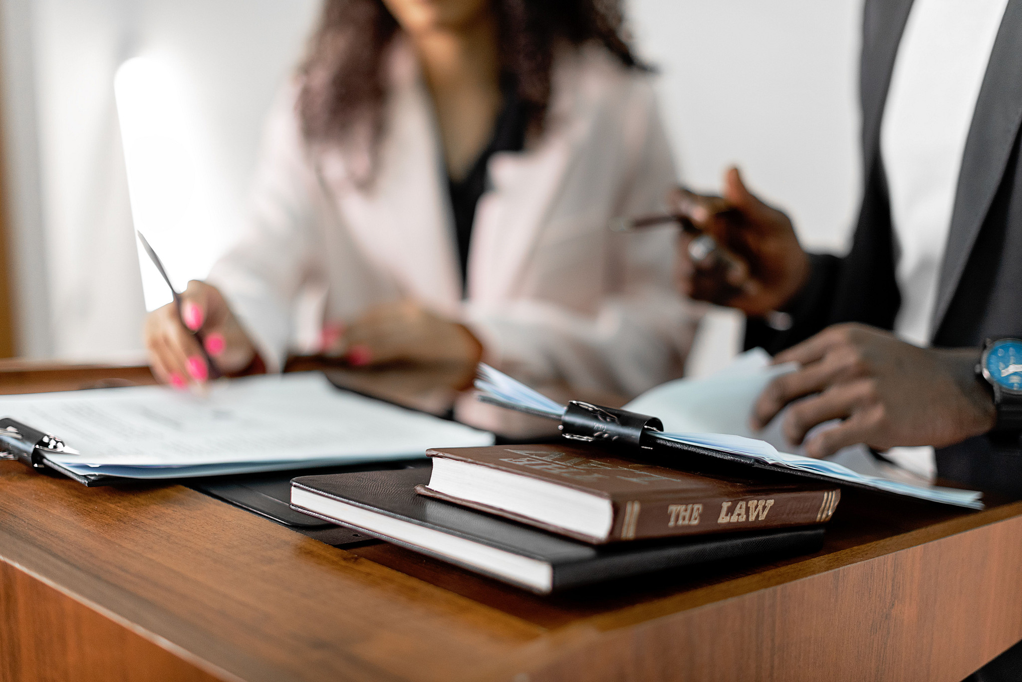 A person in a white blazer and brown hair and a person of color sit at a desk with a law book and papers in front of them
